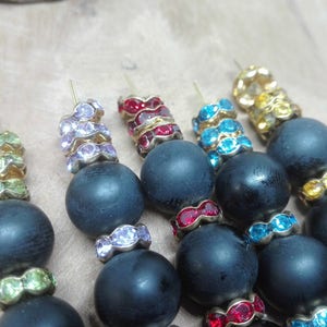Può includere: Primo piano di diversi orecchini con perline di legno blu scuro. Ogni orecchino presenta una grande perla rotonda e un accento decorativo di strass colorati nelle tonalità del verde, viola, rosso, blu e oro.