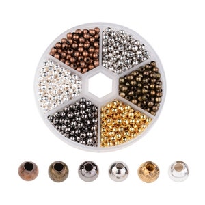 May include: A round plastic container with six compartments filled with different colored metal beads. The beads are silver, gold, bronze, copper, black, and white. There are six individual beads below the container, one of each color.