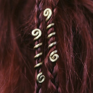 Op de afbeelding: Twee goudkleurige metalen haarclips met een spiraalvormig ontwerp worden gebruikt om een ​​gevlochten deel van donkerrood haar vast te houden.