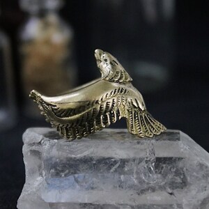 Op de afbeelding: Een ring van goudkleurig metaal met een gedetailleerd ontwerp van een vogel in vlucht. De ring wordt getoond op een helder kwartskristal.