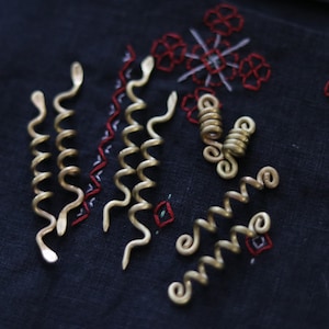 Puede incluir: Un conjunto de pinzas para el cabello de color dorado en varias formas, incluyendo espirales y bobinas. Las pinzas están dispuestas sobre un fondo de tela azul oscuro con bordados rojos y blancos.