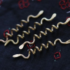 May include: Five gold-colored hair clips shaped like snakes, arranged in a row on a dark blue fabric with red and white embroidery.