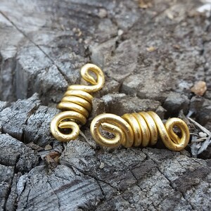 May include: Two gold-colored metal hair beads, also known as loc jewelry, are shown on a rough, gray, wooden surface. The beads are coiled and have a smooth, shiny finish.