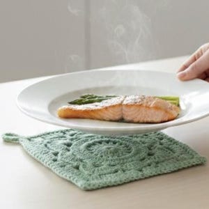 Peut inclure: Une assiette blanche contenant un filet de saumon cuit et des asperges, avec de la vapeur qui s'élève. L'assiette est posée sur un dessous-de-plat carré en crochet vert clair avec un motif décoratif.