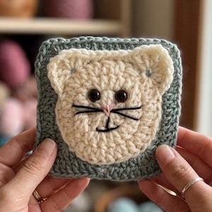 May include: A handmade crochet coaster featuring a cat face design. The cat's face is cream-colored with black whiskers and brown eyes, set against a square, sage green border. The coaster is held by a person.