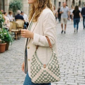 May include: A crochet shoulder bag featuring a sage green and cream geometric floral pattern. The bag has a brown leather strap attached with metal hardware and a small brown leather tab closure at the top center of the bag opening.