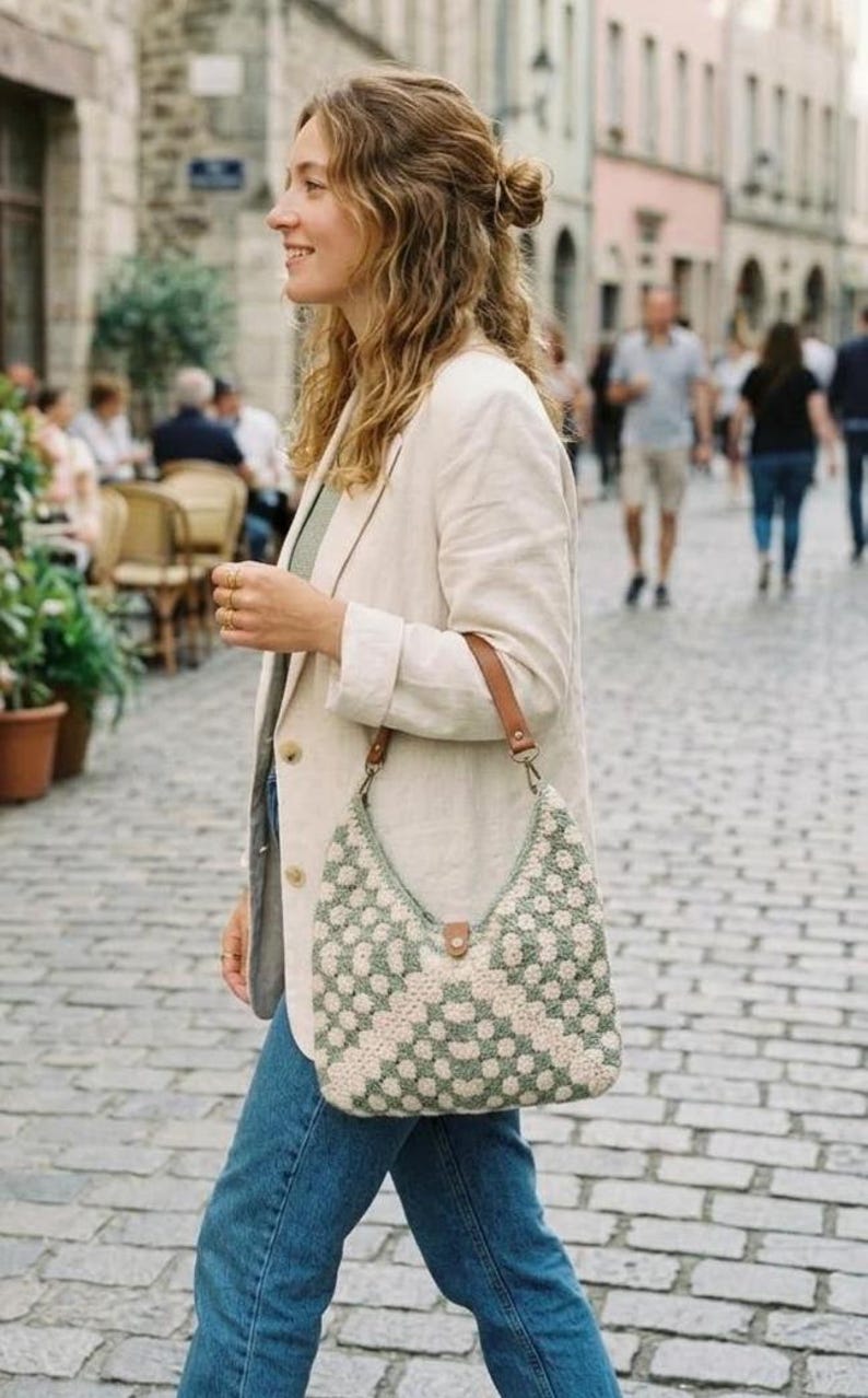 May include: A crochet shoulder bag featuring a sage green and cream geometric floral pattern. The bag has a brown leather strap attached with metal hardware and a small brown leather tab closure at the top center of the bag opening.