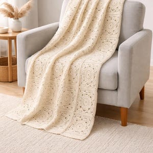 May include: Cream-colored crochet blanket draped over a gray armchair. The blanket features a repeating circular pattern within square blocks. The chair has wooden legs and sits on a cream-colored rug. A side table and vase are visible in the background.
