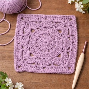 May include: A square, lilac-colored crochet piece with a detailed floral design in the center. A ball of matching yarn and a crochet hook with a wooden handle are also visible, suggesting a crafting project. The piece is on a wooden surface.