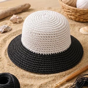 Puede incluir: Un sombrero de ganchillo de dos tonos con una corona blanca y un ala negra descansa sobre arena. El sombrero está rodeado de materiales de artesanía, incluyendo hilo y un ganchillo. También se ven conchas marinas y madera flotante.