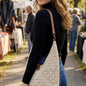 May include: A beige crochet shoulder bag with a triangular shape and a brown leather strap. The bag features a textured, woven pattern and is attached to the strap with a metal clasp and a brown leather tab detail at the top corner.