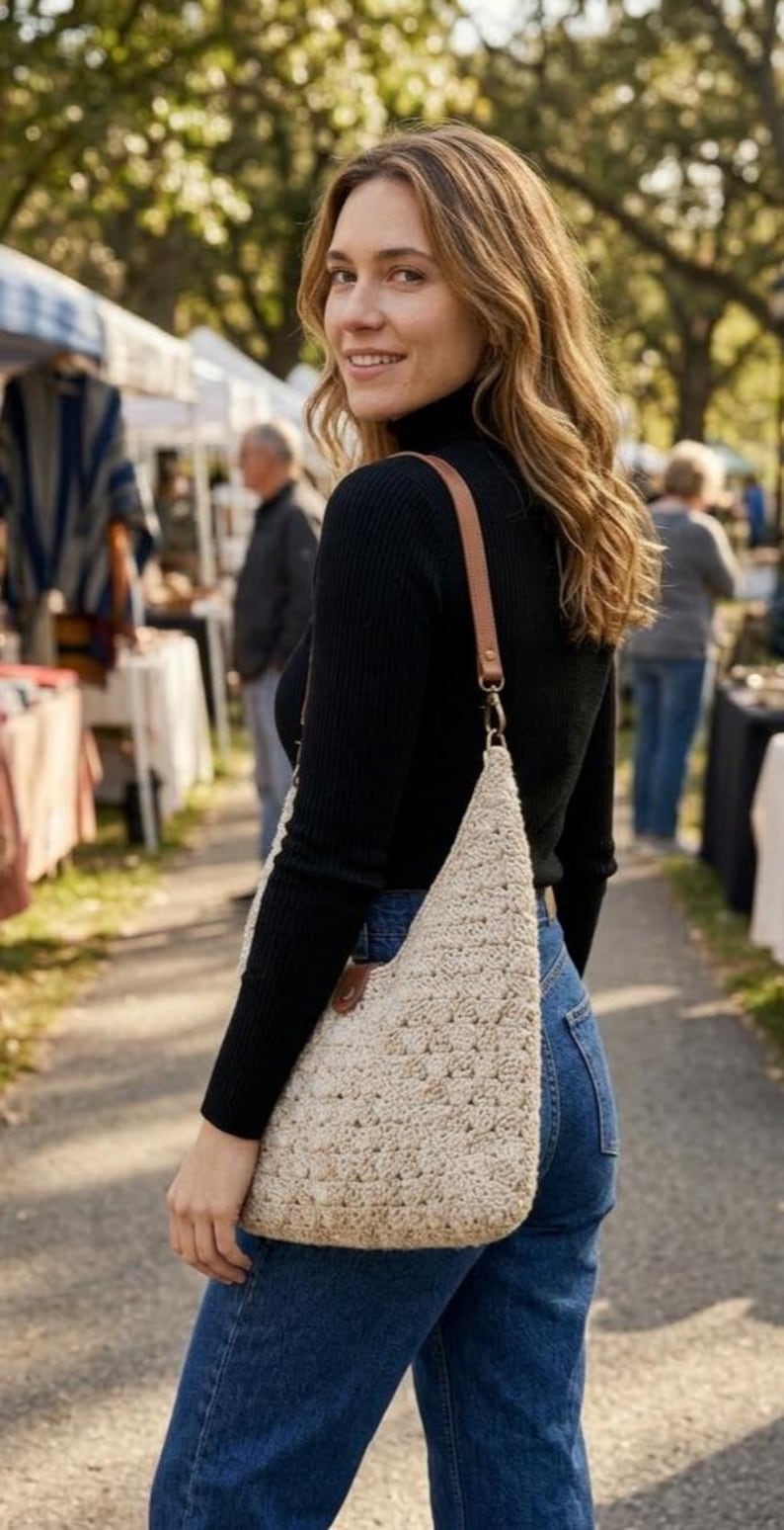 May include: A beige crochet shoulder bag with a triangular shape and a brown leather strap. The bag features a textured, woven pattern and is attached to the strap with a metal clasp and a brown leather tab detail at the top corner.
