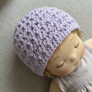 May include: A light purple crochet beanie with a textured, repeating stitch pattern worn by a fabric doll with closed eyes, a soft beige complexion, and a light-colored dress. The hat features a dense, woven construction.