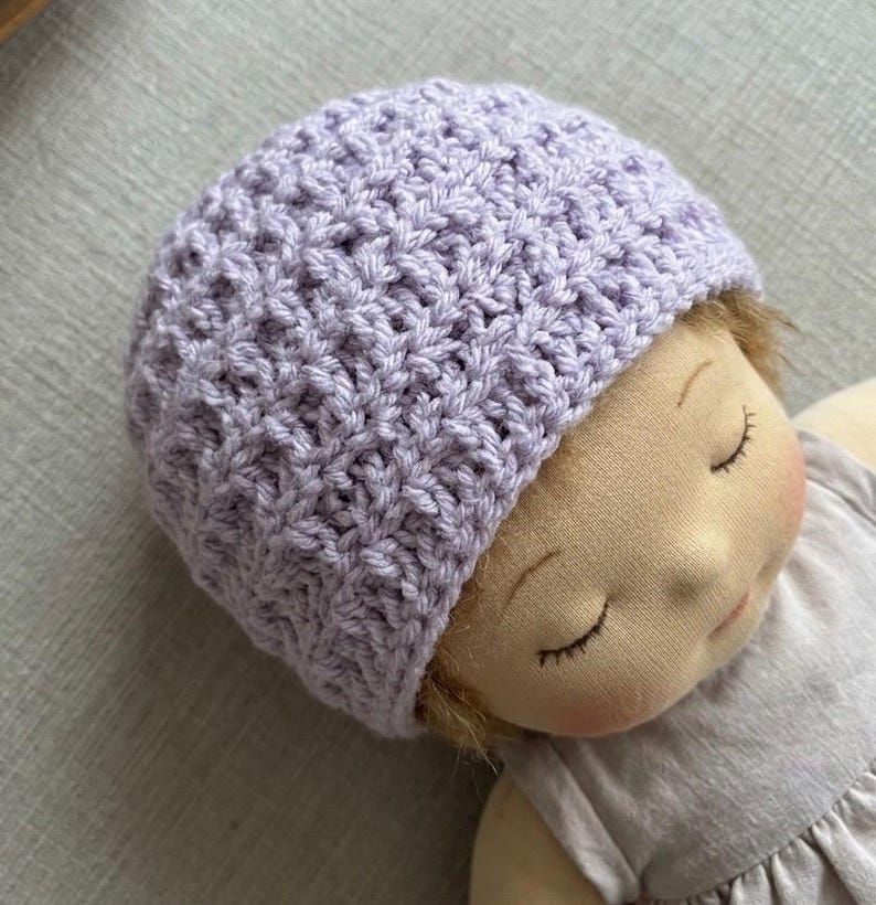 May include: A light purple crochet beanie with a textured, repeating stitch pattern worn by a fabric doll with closed eyes, a soft beige complexion, and a light-colored dress. The hat features a dense, woven construction.