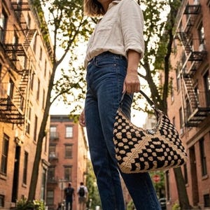 May include: A beige button-down shirt, dark blue denim jeans with a raw hem, and brown leather thong sandals. A black and beige crocheted hobo bag with a geometric pattern is held by the handle, featuring a central silver-toned metal clasp.