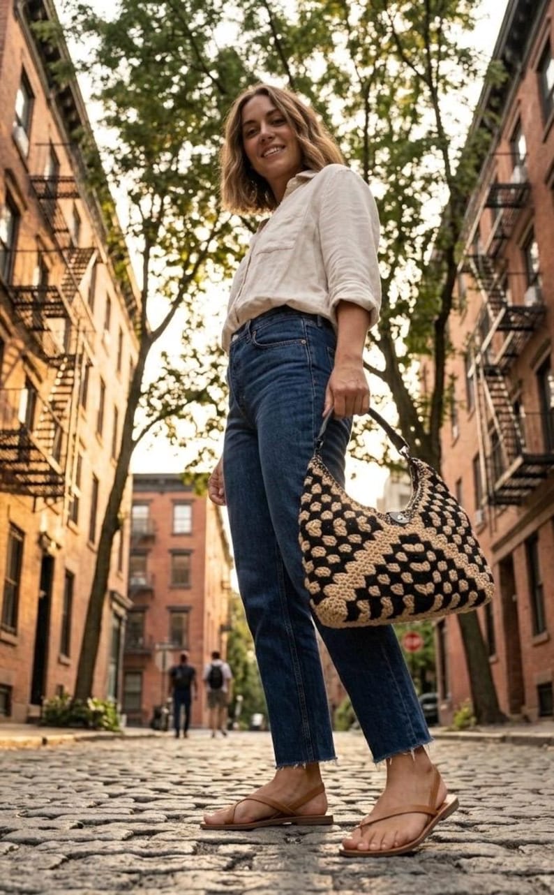 May include: A beige button-down shirt, dark blue denim jeans with a raw hem, and brown leather thong sandals. A black and beige crocheted hobo bag with a geometric pattern is held by the handle, featuring a central silver-toned metal clasp.