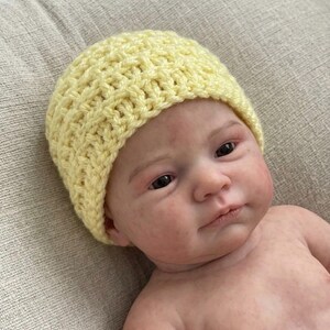 May include: A pale yellow, hand-crocheted beanie hat with a textured, waffle-like stitch pattern. The hat is worn by a realistic-looking infant doll with fair skin, dark eyes, and subtle facial features, showcasing the knit construction.