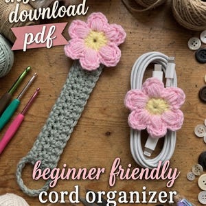 May include: A crochet pattern for a cord organizer. The image shows a gray crochet cord wrap with a pink and yellow flower, and a white cord with a similar flower. Text reads "instant download pdf" and "beginner friendly cord organizer crochet pattern."