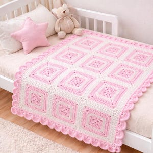 May include: A handmade, pink and white crochet blanket with a scalloped edge, displayed on a white cot. The blanket features a repeating square pattern. A pink star-shaped cushion and a teddy bear are also on the bed.