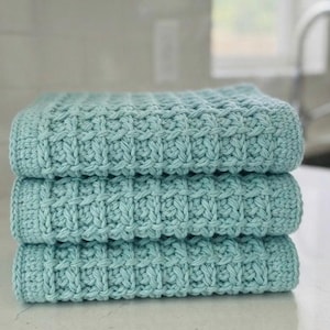 May include: A stack of three light teal hand towels featuring a textured waffle stitch pattern. Each towel is folded into a neat rectangle, showcasing the consistent grid-like design and the soft, matte finish of the yarn used in the construction.