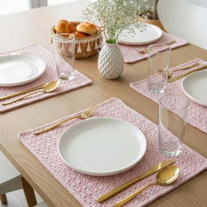 Könnte beinhalten: Ein gedeckter Esstisch mit rosa gehäkelten Tischsets, weißen Tellern, goldenem Besteck und klaren Gläsern. Ein Brotkorb und eine weiße Vase mit Blumen vervollständigen die Szene.