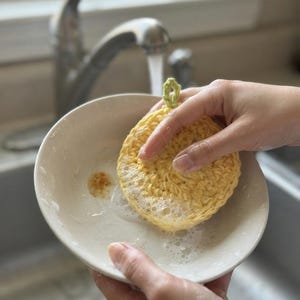 May include: A round, yellow, crocheted dish scrubber with a small green loop at the top, being used to clean a white ceramic bowl with soapy water. The textured, circular yarn scrubber features a dense stitch pattern and is covered in bubbles.