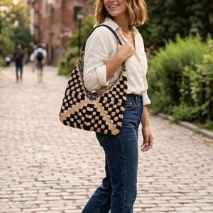 May include: A crochet shoulder bag featuring a black and beige checkered pattern, a black leather strap, and a central button closure. The bag is worn over the shoulder, showcasing its textured, handcrafted design and spacious hobo-style shape.