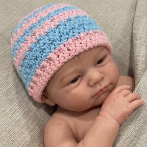 May include: A hand-crocheted beanie hat featuring alternating horizontal stripes of light pink and light blue yarn. The hat has a textured stitch pattern and a ribbed edge, worn by a realistic doll with light skin and brown hair.