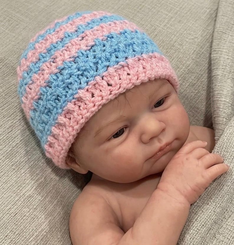 May include: A hand-crocheted beanie hat featuring alternating horizontal stripes of light pink and light blue yarn. The hat has a textured stitch pattern and a ribbed edge, worn by a realistic doll with light skin and brown hair.