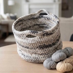 May include: A handmade crochet basket with a striped pattern in shades of gray, beige, and white. The basket has two handles and is accompanied by several balls of yarn in complementary colors, suggesting a craft or storage theme.