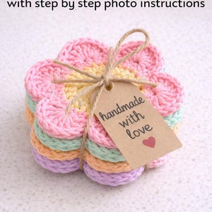 May include: Stack of colorful crochet flower coasters tied with twine and a tag that reads "handmade with love". The coasters are in shades of pink, yellow, green, orange, and purple. The image also includes the text "CROCHET PATTERN with step by step photo instructions".