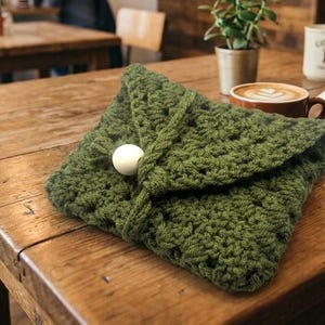 May include: A handmade, olive green crocheted pouch with a white button closure. The pouch is folded over and sits on a wooden table. A cup of coffee, a mug, and a small plant are in the background.