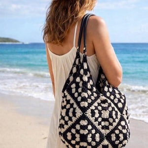 May include: A black and cream crochet tote bag featuring a repeating granny square pattern. The bag has two dark leather-like shoulder straps attached with silver-toned metal hardware, providing a structured contrast to the soft textile body.