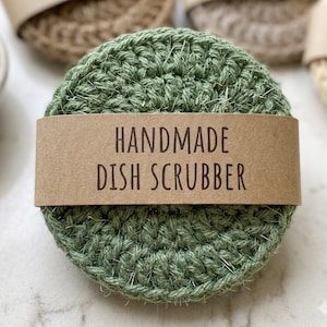 May include: A handmade, round, green crocheted dish scrubber with a brown paper band that reads "HANDMADE DISH SCRUBBER". The scrubber is made of a textured, woven material. Other scrubbers are visible in the background.