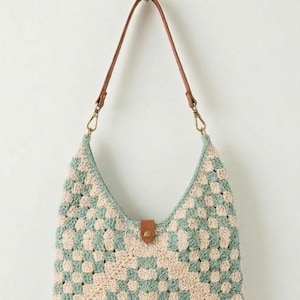 May include: A crochet hobo bag featuring a checkered pattern in sage green and cream yarn. The bag includes a brown leather strap attached with gold-toned metal clasps and a central brown leather tab closure with a gold-toned metal snap button.