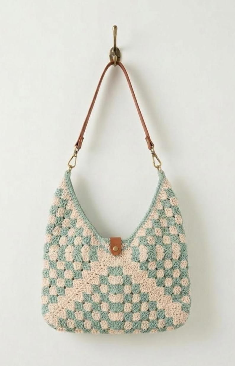 May include: A crochet hobo bag featuring a checkered pattern in sage green and cream yarn. The bag includes a brown leather strap attached with gold-toned metal clasps and a central brown leather tab closure with a gold-toned metal snap button.