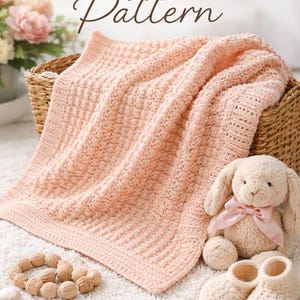 May include: A peach-colored crochet blanket with a textured pattern rests in a wicker basket. A stuffed bunny with a pink bow, baby shoes, and a wooden teething ring are nearby. The image includes the text "CROCHET Pattern".