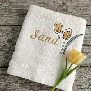 Op de afbeelding: Witte badhanddoek met geborduurd bloemmotief en de naam "Sana" in gouden letters. Een gele tulp ligt op de handdoek. De handdoek heeft een decoratieve rand.