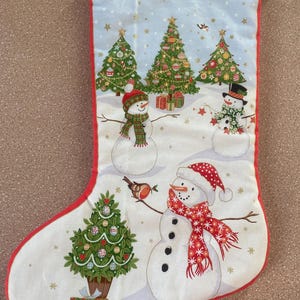 May include: Christmas stocking with a red trim and a white background. The stocking features snowmen, Christmas trees, and a robin. The snowmen wear hats and scarves. The trees are decorated with ornaments and lights. A festive holiday decoration.
