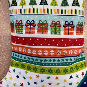 May include: A white stocking with a Christmas pattern featuring green trees, red and white stripes, and red and green gift boxes. The stocking has a white background with a repeating pattern of stars.