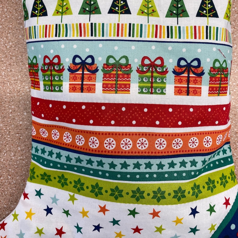 May include: A white stocking with a Christmas pattern featuring green trees, red and white stripes, and red and green gift boxes. The stocking has a white background with a repeating pattern of stars.