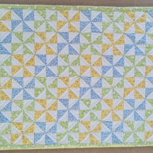 以下が含まれることがあります： 青、緑、黄色の正方形のパッチワーク模様のベビーキルト。キルトは綿生地で作られており、白い背景があります。