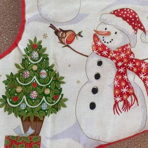 May include: A Christmas-themed fabric with a white background featuring snowmen, a Christmas tree, and a bird. The snowmen have red and white hats and scarves. The Christmas tree is decorated with ornaments and hearts. A red border frames the design.