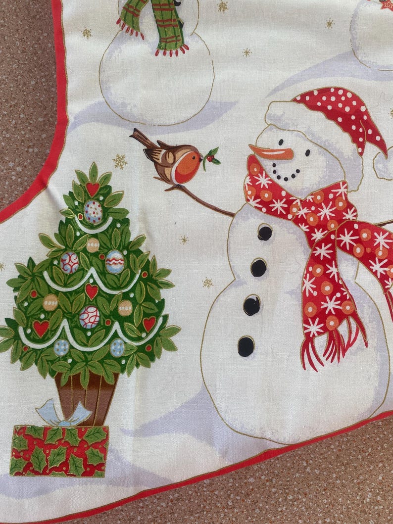 May include: A Christmas-themed fabric with a white background featuring snowmen, a Christmas tree, and a bird. The snowmen have red and white hats and scarves. The Christmas tree is decorated with ornaments and hearts. A red border frames the design.