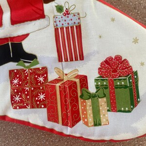 May include: A Christmas tree skirt featuring a white background with illustrations of wrapped gifts in red, green, and gold. The gifts are decorated with bows and patterns. The skirt has a red border and gold accents.
