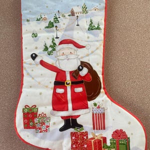 May include: Christmas stocking with a red trim and a white background. Santa Claus is featured in the center, holding a sack of gifts. The background depicts a snowy village scene with presents at the bottom.