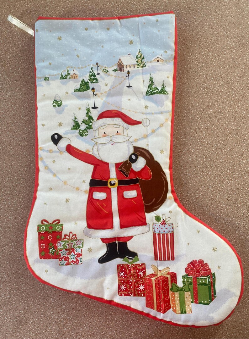 May include: Christmas stocking with a red trim and a white background. Santa Claus is featured in the center, holding a sack of gifts. The background depicts a snowy village scene with presents at the bottom.