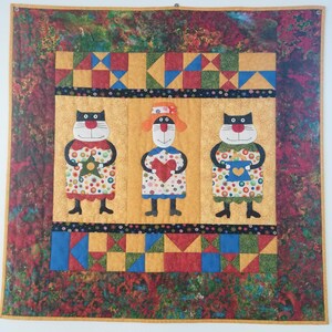 以下が含まれることがあります： パッチワークのデザインが施されたカラフルなキルトで、3匹の黒い猫がそれぞれ異なるものを抱えています。猫はドレスを着ていて、赤い鼻をしています。キルトは黄色い背景に赤、緑、青のアクセントが施されています。