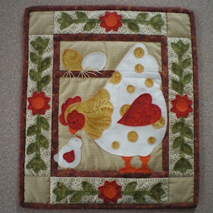 以下が含まれることがあります： 白い鶏が赤いハートと金のアクセントで描かれたキルトの壁掛け。小さなひよこ、茶色、緑、赤の花の縁取りが施されています。