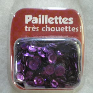May include: A clear plastic container with a red lid and the text "Paillettes très chouettes!"  The container holds a large quantity of purple sequins.